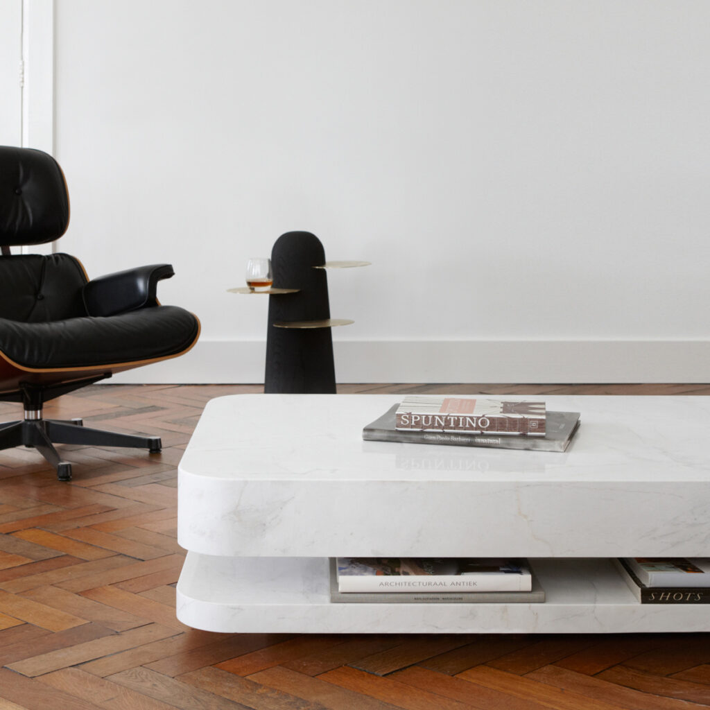 Table basse en marbre blanc à deux plateaux, des livres glissés entre eux, près d'un fauteuil en cuir noir